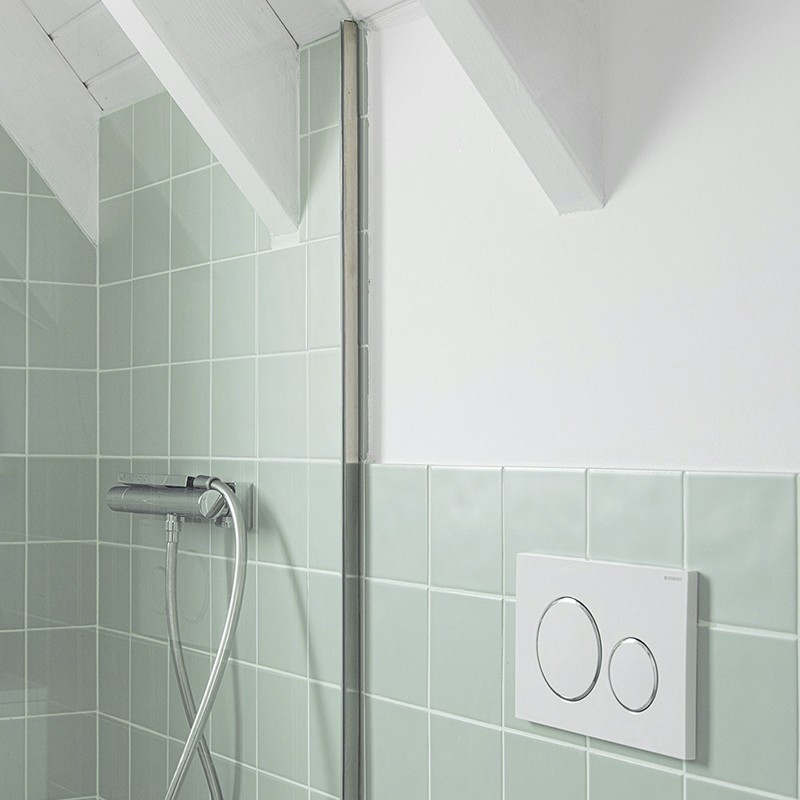Pastellgrüne quadratische Wandfliesen in Dusche; verchromte Duscharmatur mit flexibler Handbrause, sichtbare vertikale Rohrführung, weiße Wand und Betätigungsplatte mit zwei runden Tasten.