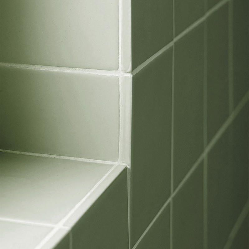 Azulejo cerámico blanco verdoso en esquina con rejuntes visibles; forma un escalón o repisa, y la pared contigua muestra una cuadrícula de azulejos alineados.
