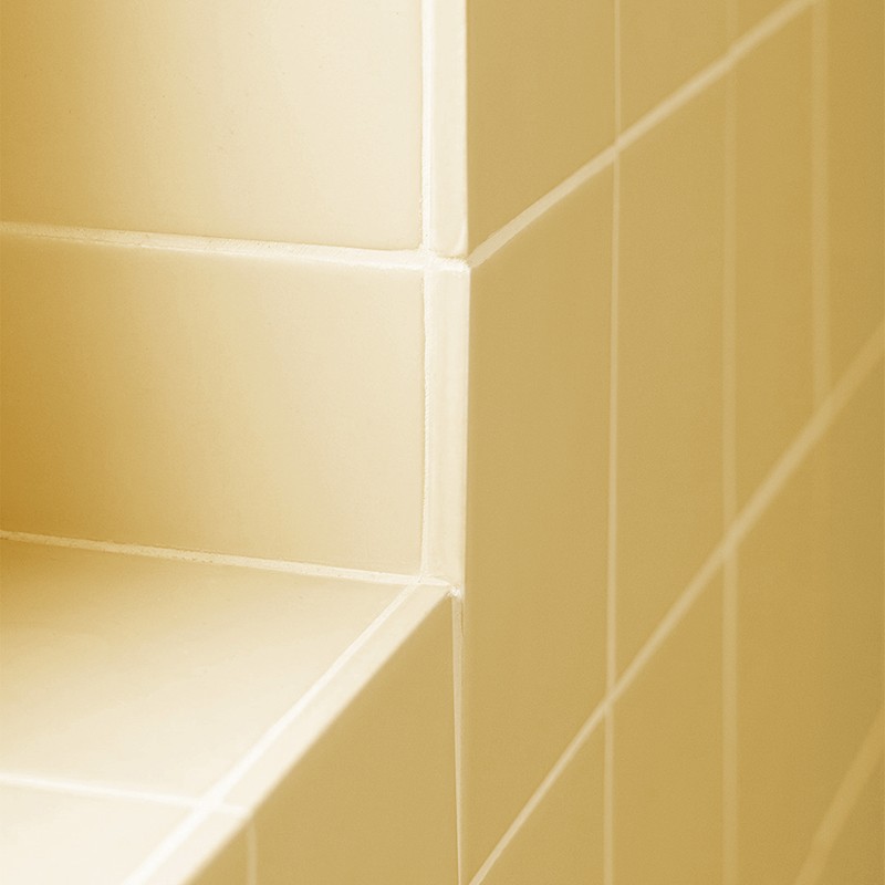 Azulejos cerâmicos bege-claro formando um canto com prateleira embutida; juntas brancas visíveis e acabamento brilhante, aparência limpa e minimalista.