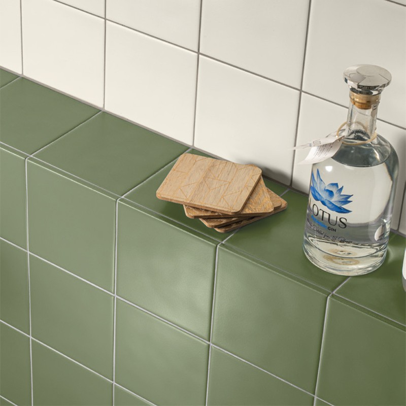 Azulejo verde mate rectangular formando una repisa frente a azulejos blancos; sobre él hay posavasos de madera y una botella con etiqueta azul.   Texto visible: "LOTUS". Texto pequeño en la etiqueta y en la etiqueta colgante no legible.