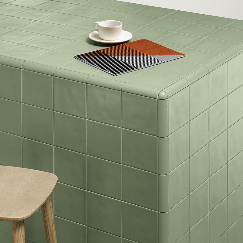 Azulejo verde mate cuadrado formando una encimera curva; sobre ella, taza blanca con platillo y una muestra geométrica, taburete de madera junto al frente.