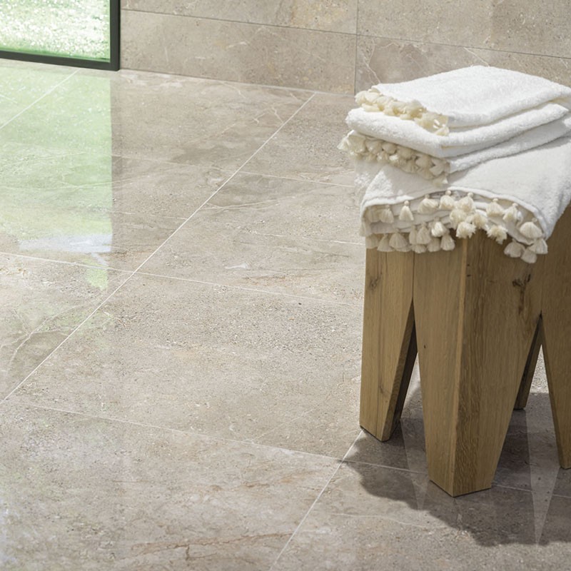 Baldosa cerámica pulida beige con vetas claras; suelo junto a un ventanal, acompañado de un pequeño taburete de madera con toallas blancas con borlas.