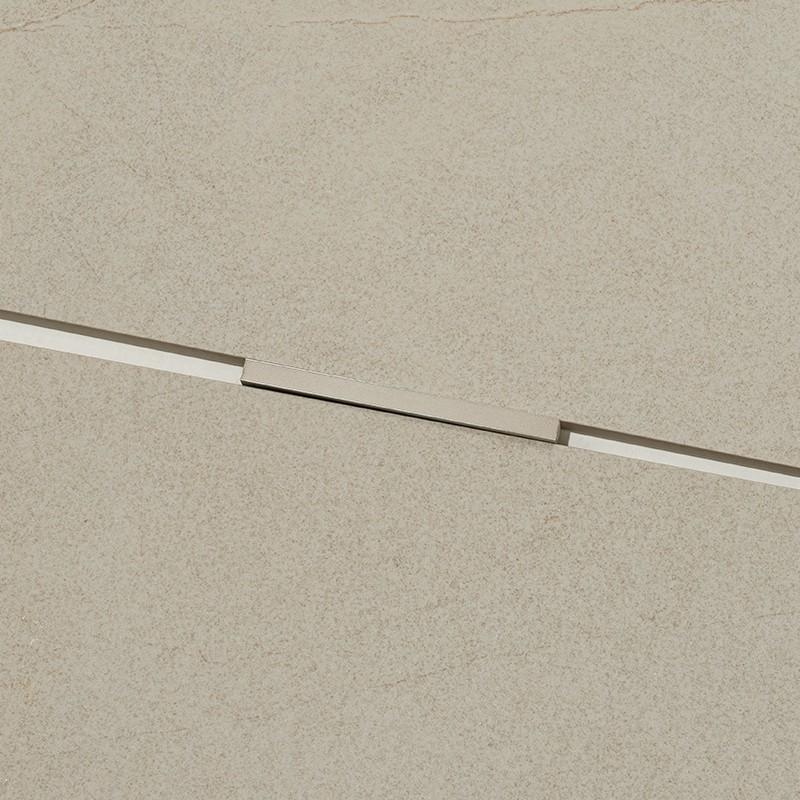 Azulejo beige texturizado con ranura diagonal estrecha y tapa metálica rectangular ligeramente elevada; contexto: superficie continua de suelo o pared del mismo tono, sin otros elementos visibles.