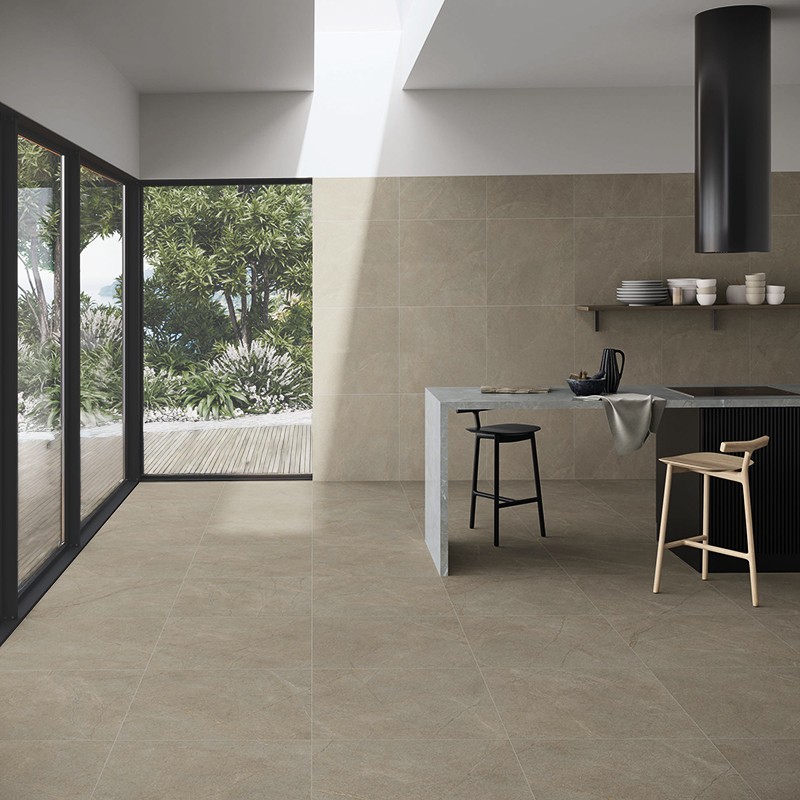Square matte beige tiles on floor and wall; minimalist open kitchen with gray island, stools, shelving, large sliding window and overhead light illuminating the exterior garden.