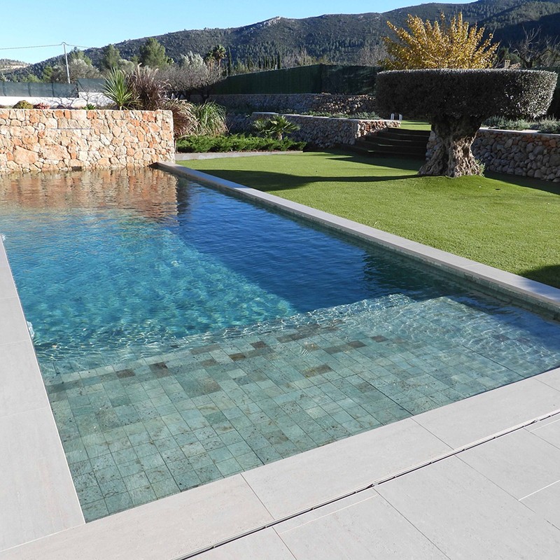 Quadratische grünlich-blaue Fliesen auf Poolboden; klares Wasser mit sichtbarer Stufenkante, helle Beckenrandplatten; Umgebung: Kunstrasen, niedrige Natursteinmauer, Buschwerk und Hügel im Hintergrund.