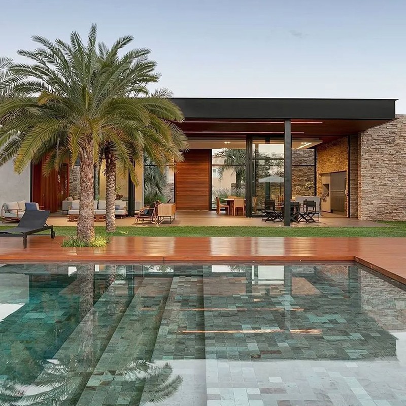 Carrelage de piscine en mosaïque vert-bleu (bassin peu profond), devant terrasse en bois, palmiers, mobilier extérieur et façade vitrée moderne avec mur en pierre.