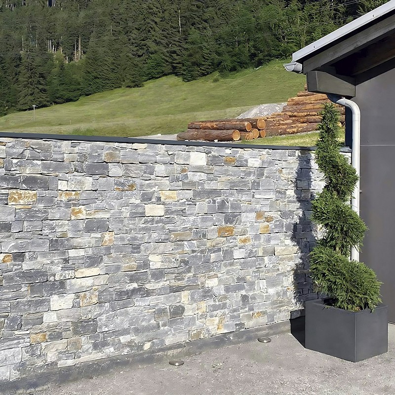 Mur de parement en pierres grises en premier plan; à droite pot carré avec buisson taillé, derrière pelouse en pente, tas de rondins empilés et lisière de forêt.