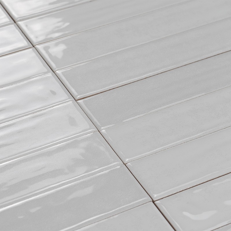 Azulejos retangulares brancos e brilhantes, assentados em fileiras com juntas visíveis, superfície levemente ondulada refletindo luz, sugerindo parede ou piso interior.