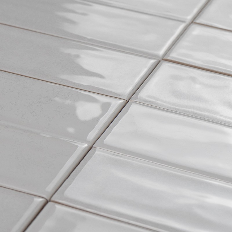 Carrelage rectangulaire blanc brillant, bords légèrement irréguliers et joints fins gris clair; plan rapproché montrant reflets et texture émaillée.