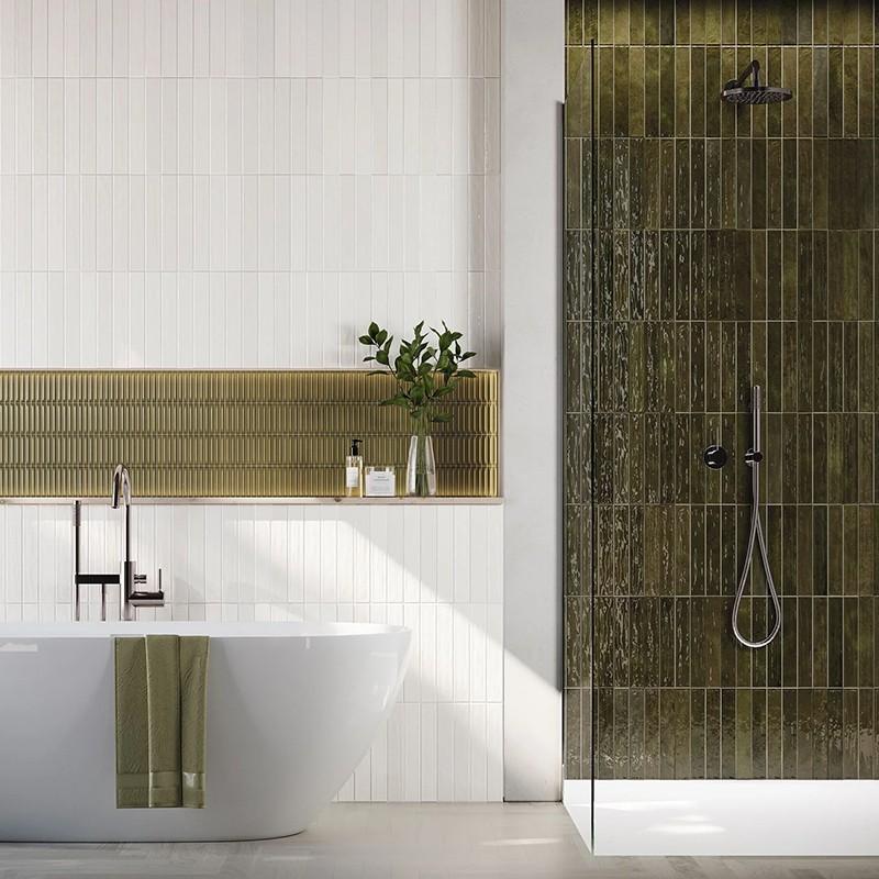 Groene verticale geglazuurde tegels bedekken een douchewand met zwart mat douchebeslag, naast witte wandtegels, vrijstaand wit bad met groene handdoek en plank met planten en toiletartikelen.