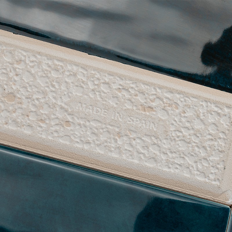 Azulejo rectangular crema con textura y texto en relieve "MADE IN SPAIN"; rodeado por azulejos esmaltados azul oscuro con reflejos y juntas estrechas.