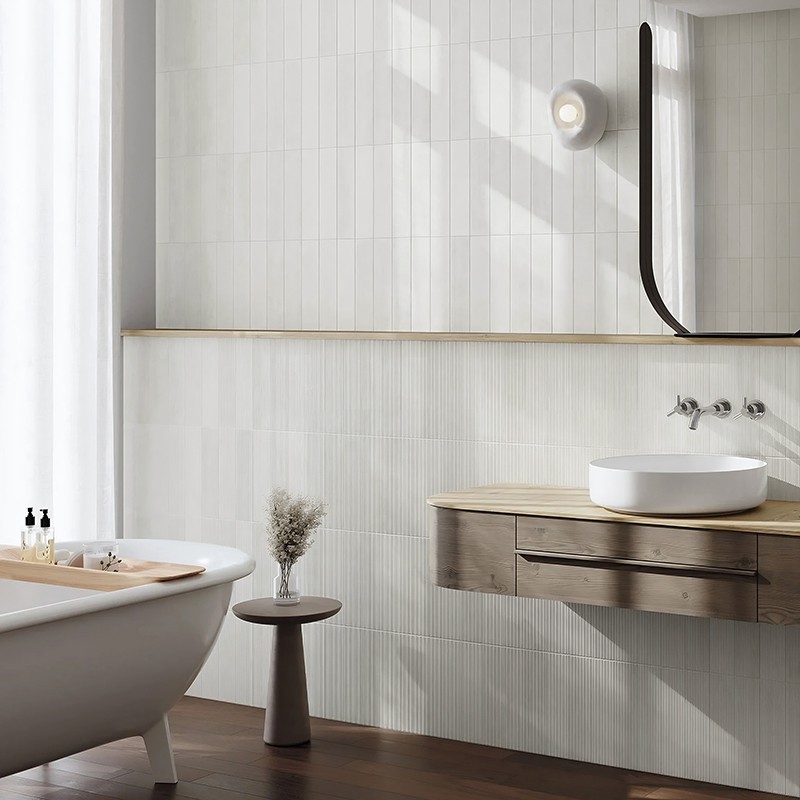 Lichte wandtegel met subtiele verticale ribbelstructuur; moderne badkamer met vrijstaand wit bad, houten wastafelmeubel en ronde witte kom, donker houten vloer en zachte daglichtinval.