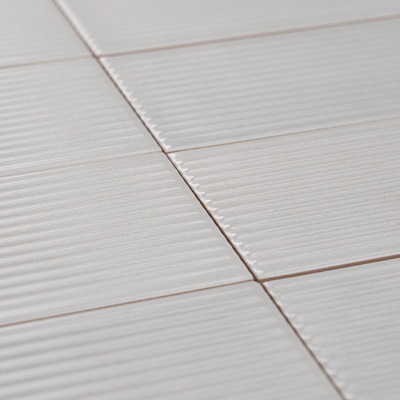 Azulejos brancos retangulares com superfície estriada, rejunte fino creme entre peças, dispostos em padrão regular sobre parede ou piso.