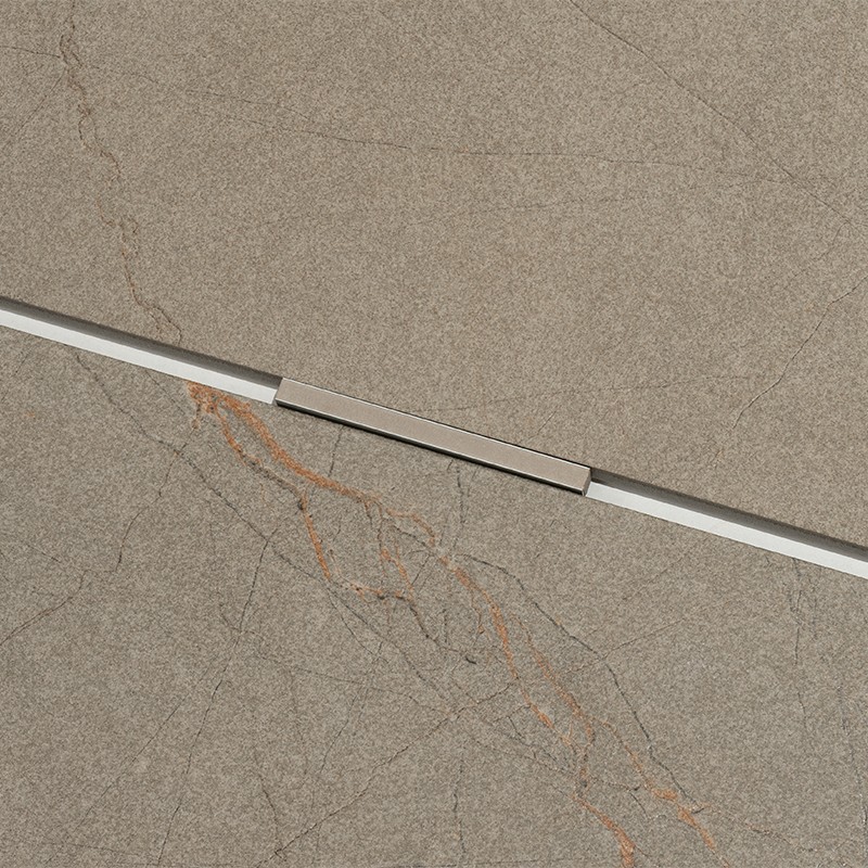 Azulejo cinza-amarronzado com veios finos acastanhados; junta diagonal com perfil metálico prateado e conector retangular central, superfície texturizada tipo pedra.