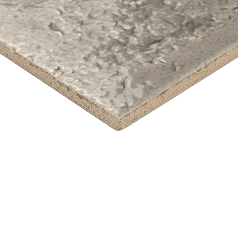 Azulejo cerámico con superficie esmaltada y textura irregular grisácea; se muestra el canto y el núcleo sin esmaltar, en primer plano sobre fondo blanco.