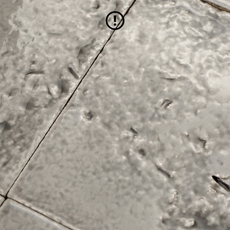 Off-white glazed tile, irregular texture with small cracks and marks; dark diagonal joints; a circle with '!' appears in the upper corner.