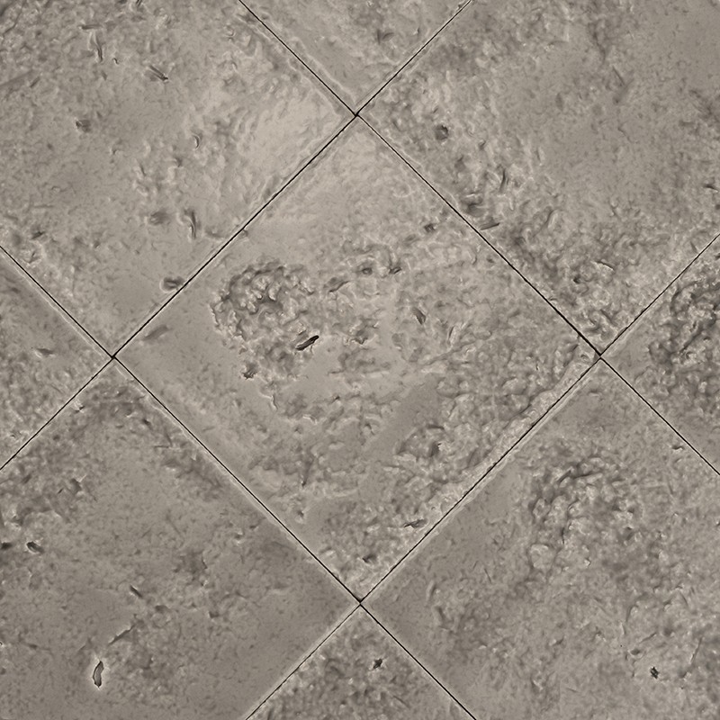 Baldosa cerámica gris claro, textura moteada y relieve irregular; juntas en cruz forman patrón diagonal con baldosas similares alrededor, aspecto ligeramente desgastado y tonos moteados.