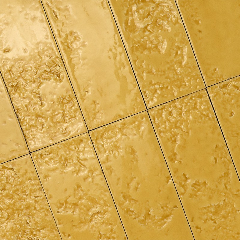 Carrelage rectangulaire jaune doré, surfaces brillantes et bosselées; joints fins et sombres forment une grille diagonale, évoquant un sol ou un mur carrelé.