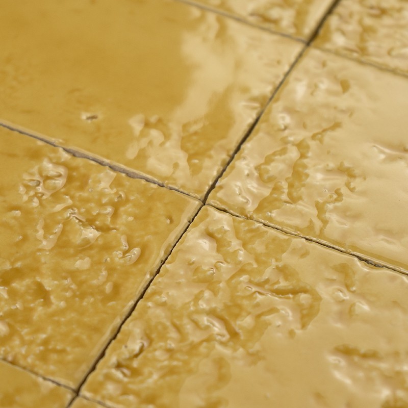 Azulejo cerâmico amarelo com superfície irregular e brilhante, rejunte escuro formando cruz; close-up mostrando água ou brilho refletido, sugerindo piso molhado.
