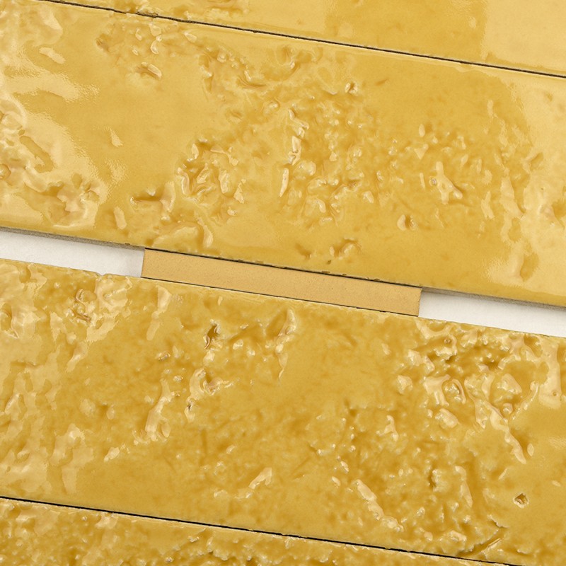 Rectangular ceramic tile in mustard color with a rough texture and glazed shine; arranged in horizontal rows with thin, clear joints between pieces.