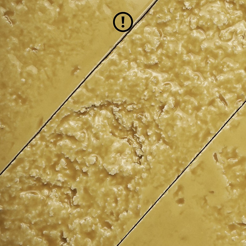 Fliese mit rauer, abgenutzter Mittelrille, zwei diagonalen Fugen, gelb-beige Oberfläche; oben ein kleines Ausrufezeichen in einem Kreis ("!").