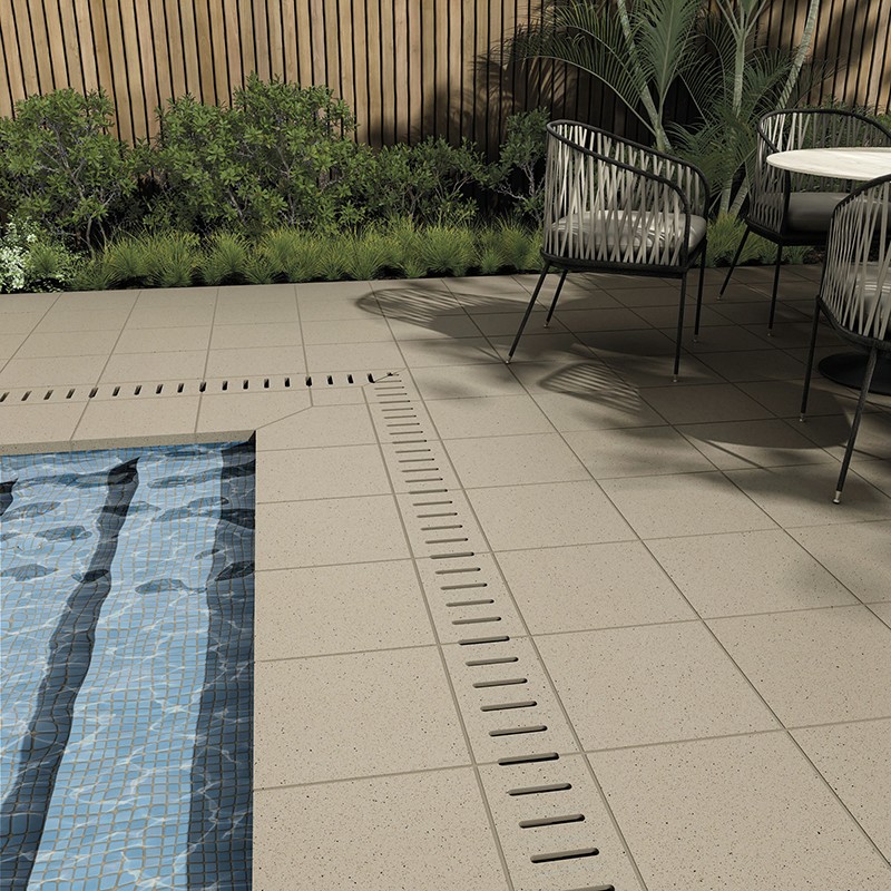 Baldosa gris texturada junto a desagüe lineal, borde de piscina con mosaico azul, sillas de jardín, plantas y valla de madera al fondo.