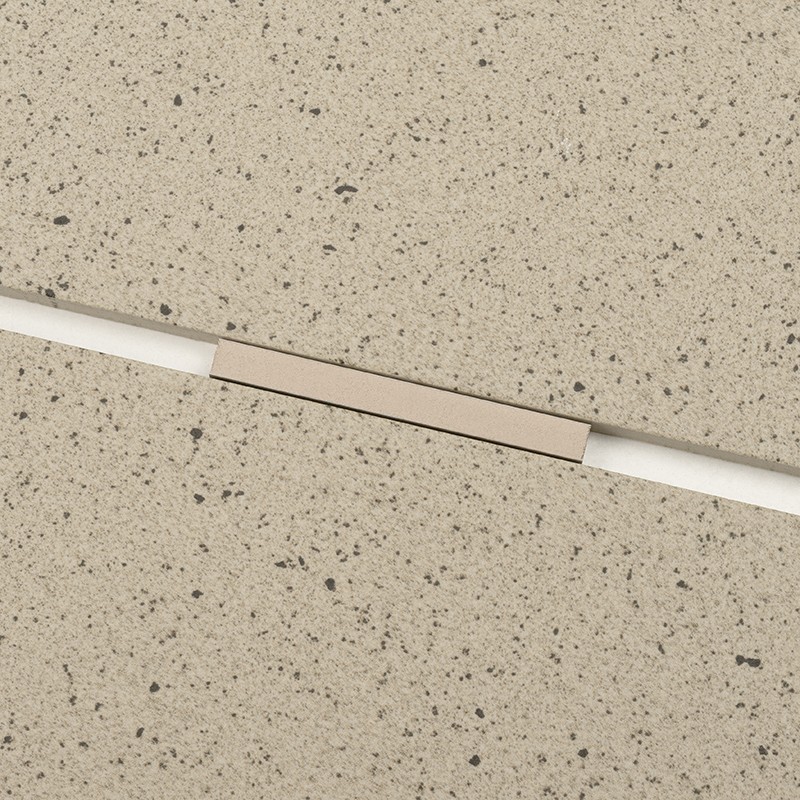 Azulejo beige claro con motas oscuras; junta diagonal blanca atraviesa la superficie y un pequeño tramo rectangular beige metálico cubre la unión.