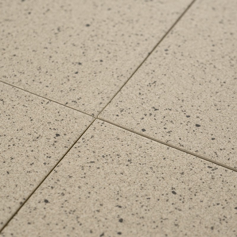 Azulejo beige salpicado de motas oscuras; cuatro baldosas cuadradas unidas por juntas finas formando una cruz, superficie ligeramente texturada, pavimento interior neutro.