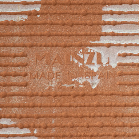 Terracotta tile sits with embossed text, `MANZU MADE IN SPAIN,` featuring horizontal ridges and white speckled areas, suggesting Terracotta tile sits with embossed text, `MANZU MADE IN SPAIN,` featuring horizontal ridges and white speckled areas, suggesting