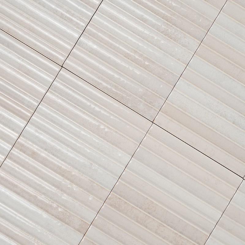 Azulejos retangulares ondulados beges dispostos em padrão modular, juntas finas escuras, superfície acetinada com variações sutis de tom e marcas horizontais; visão em ângulo diagonal.