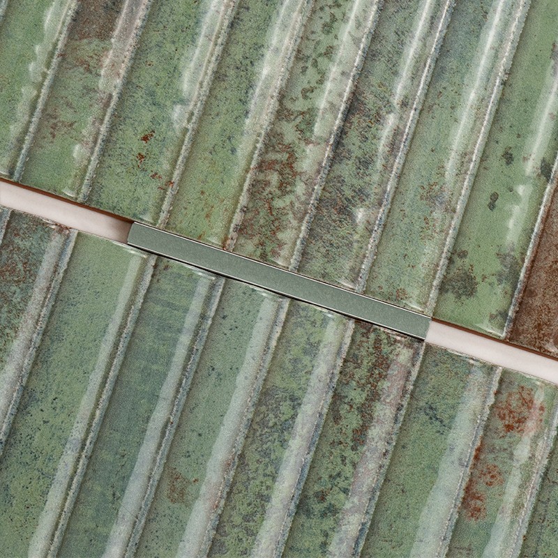 Carrelage vert rectangulaire à rainures verticales, finition usée et taches brunes; joint beige traversé en diagonale par une fine baguette métallique, surface patinée et mouchetée.