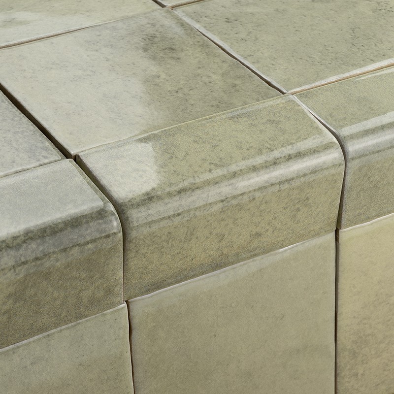 Carrelage céramique vert pâle aux angles arrondis, joints étroits; surface brillante et marbrée couvrant l'arête et la face d'un plan horizontal.