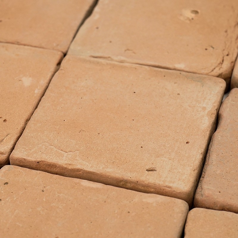 Baldosa cuadrada de terracota, superficie porosa y cantos algo irregulares; insertada entre baldosas iguales formando un pavimento con juntas estrechas.