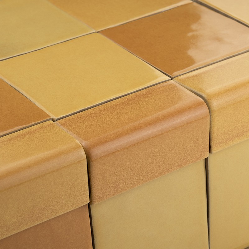 Azulejo cerámico cuadrado esmaltado en tonos ocres y beige, en esquina de superficie, con juntas visibles y bordes redondeados formando una alineación regular.