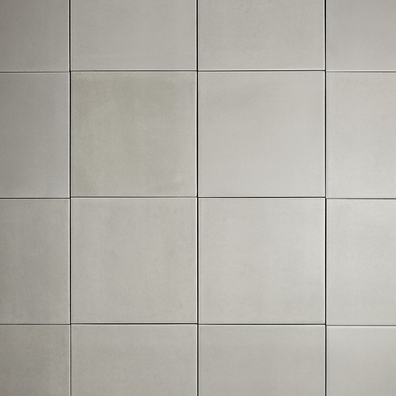 Azulejos cuadrados lisos gris claro (16) con juntas finas oscuras; patrón cuadriculado uniforme en una pared, algunos azulejos muestran leves variaciones tonales.