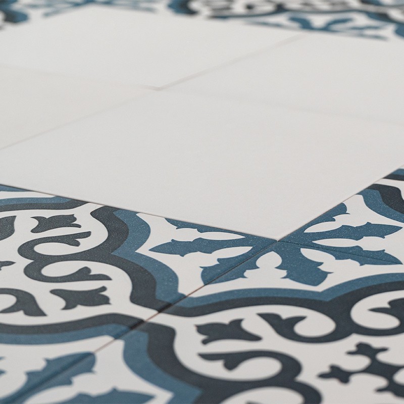 Fliese mit weißer, ungemusterter Mitte und blau-schwarzem ornamentalen Rand; Teil einer Bodenverlegung, sichtbare Fugen und angrenzende, identisch gemusterte Fliesen.