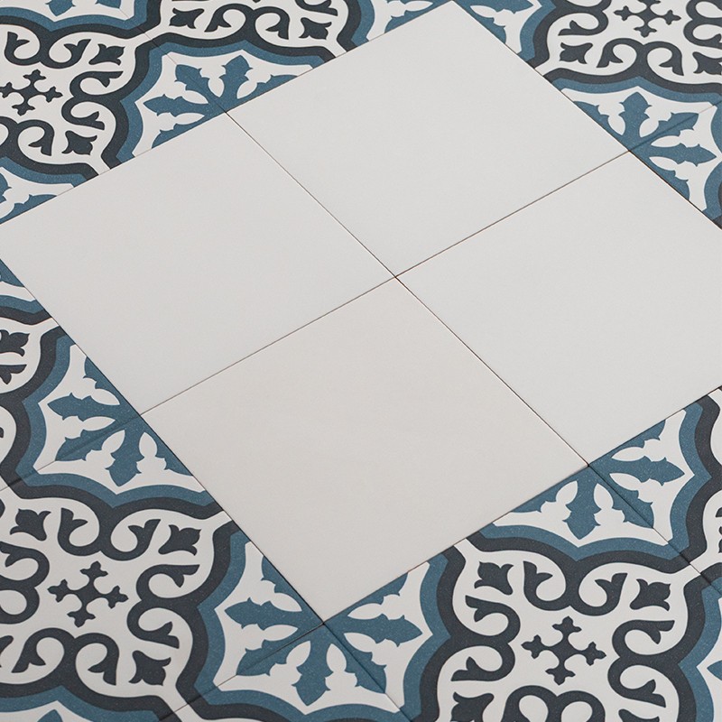 Vier weiße quadratische Fliesen bilden ein Kreuz; umgeben von blau-grauen ornamentalen Fliesen mit schwarzen, geschwungenen Konturen.