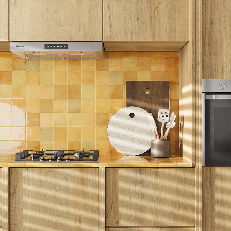 Bright yellow and orange square tile as a backsplash; setting: kitchen with wooden countertop, gas hob, extractor hood, built-in oven, boards and utensils, sunlight casting shadows.
