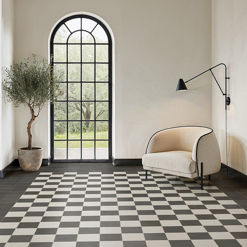 Azulejos de suelo blanco y gris en patrón ajedrezado; habitación luminosa con ventana de arco de metal, sillón beige curvo, lámpara de pared negra y maceta con árbol.