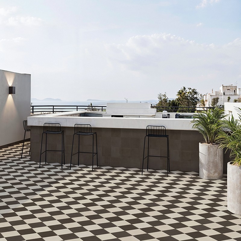 Black and white checkered pattern tile covers a wide terrace in front of a bar with three stools, a railing overlooking the sea, and large pots with palm trees.