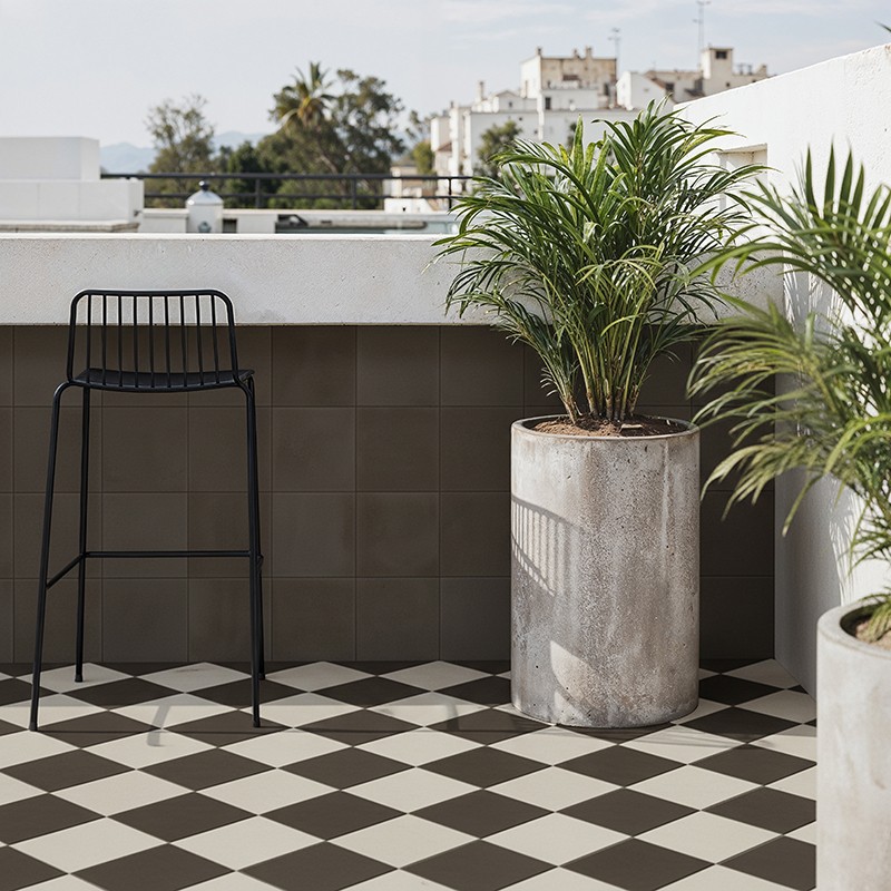 Azulejo quadriculado preto e branco no chão; varanda com parapeito branco, banco alto metálico preto e vasos de cimento com palmeiras, edifícios baixos ao fundo sob céu claro.