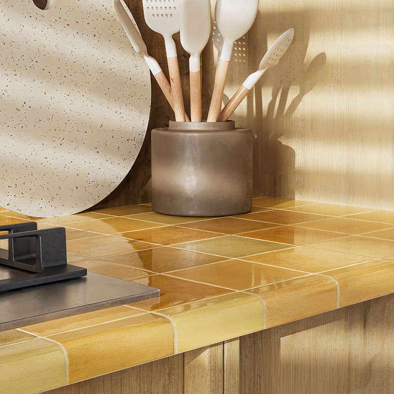 Yellow glazed tile on countertop; surroundings: stovetop, utensil holder with silicone spatulas in ceramic container, round table and warm sunlight on wooden furniture.