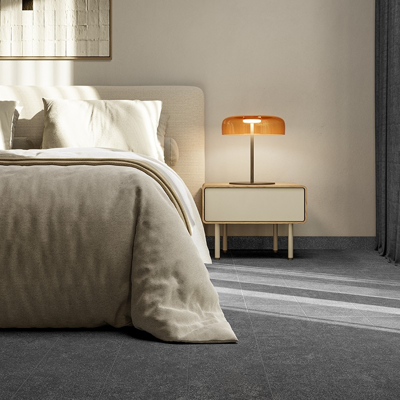 Baldosa gris mate rectangular en primer plano; dormitorio moderno con cama beige, mesita blanca y lámpara naranja sobre suelo con luz natural y cortina gris al fondo.