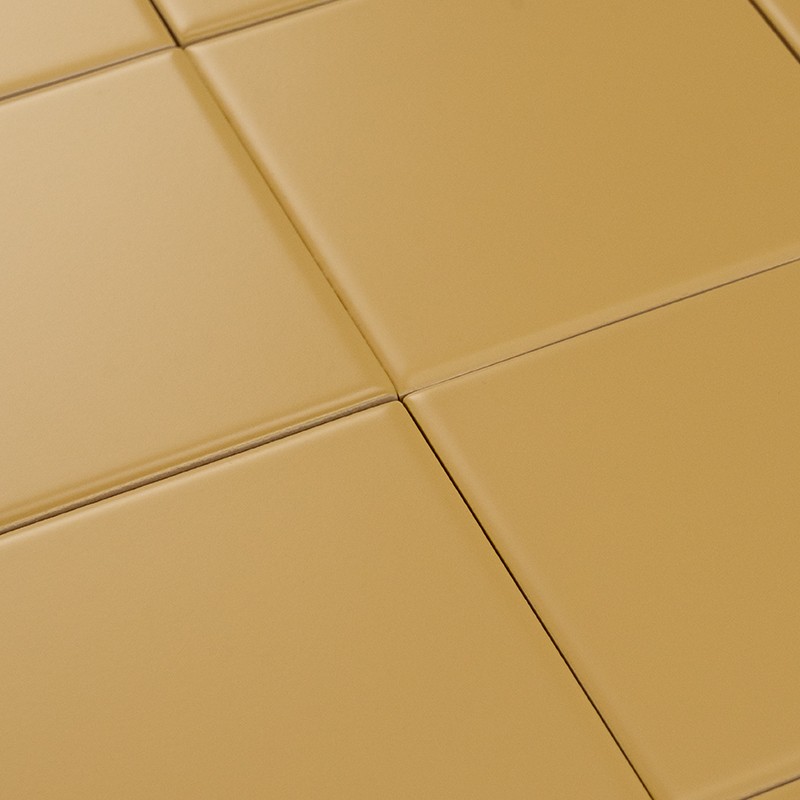Azulejos cuadrados cerámicos color mostaza, superficie lisa y mate, bordes ligeramente biselados; juntas finas crean un patrón regular sobre una superficie horizontal.