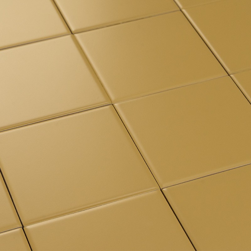 Square matte gold tiles, arranged in a grid with thin joints; smooth and slightly curved surface, illuminated by side light that creates soft shadows.