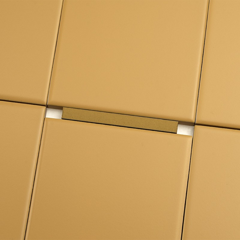 Carrelage beige à grands carreaux; joints sombres et, dans une fente entre deux carreaux, un petit insert métallique doré encadré par deux cales blanches.