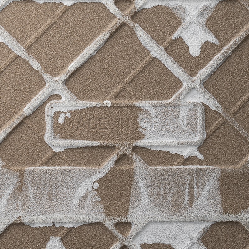 Azulejo bege texturizado com relevo geométrico; resíduos brancos de argamassa preenchendo sulcos; selo em relevo central com o texto: MADE IN SPAIN  Transcrição do texto na imagem: MADE IN SPAIN