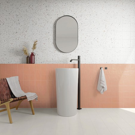 A white pedestal sink with a sleek black faucet stands against a pink and white speckled tile wall. Nearby are a mirror, a chair A white pedestal sink with a sleek black faucet stands against a pink and white speckled tile wall. Nearby are a mirror, a chair