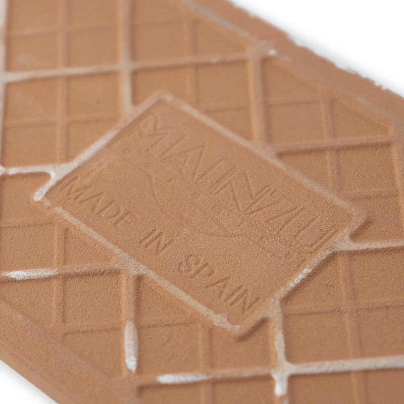 Um ladrilho retangular de cor terracota apresenta texto em relevo que diz `MANUAL` e `MADE IN SPAIN`, com um padrão de grade e a