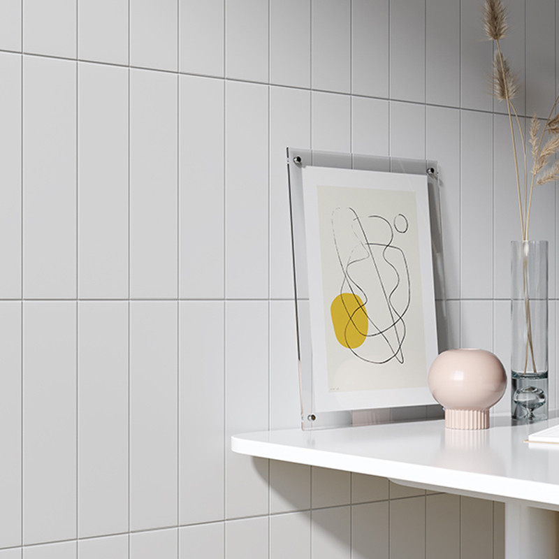 A framed abstract line drawing with a yellow circle rests on a white ledge against tiled walls, alongside a round vase and tall 